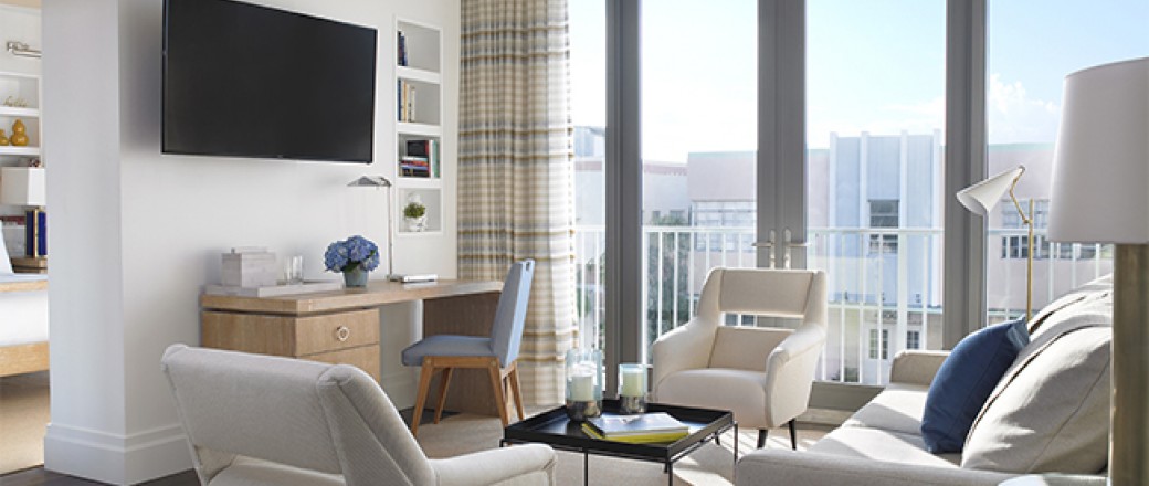 In room living space with beige couches, black coffee table, wooden desk with chair, Tv & balcony